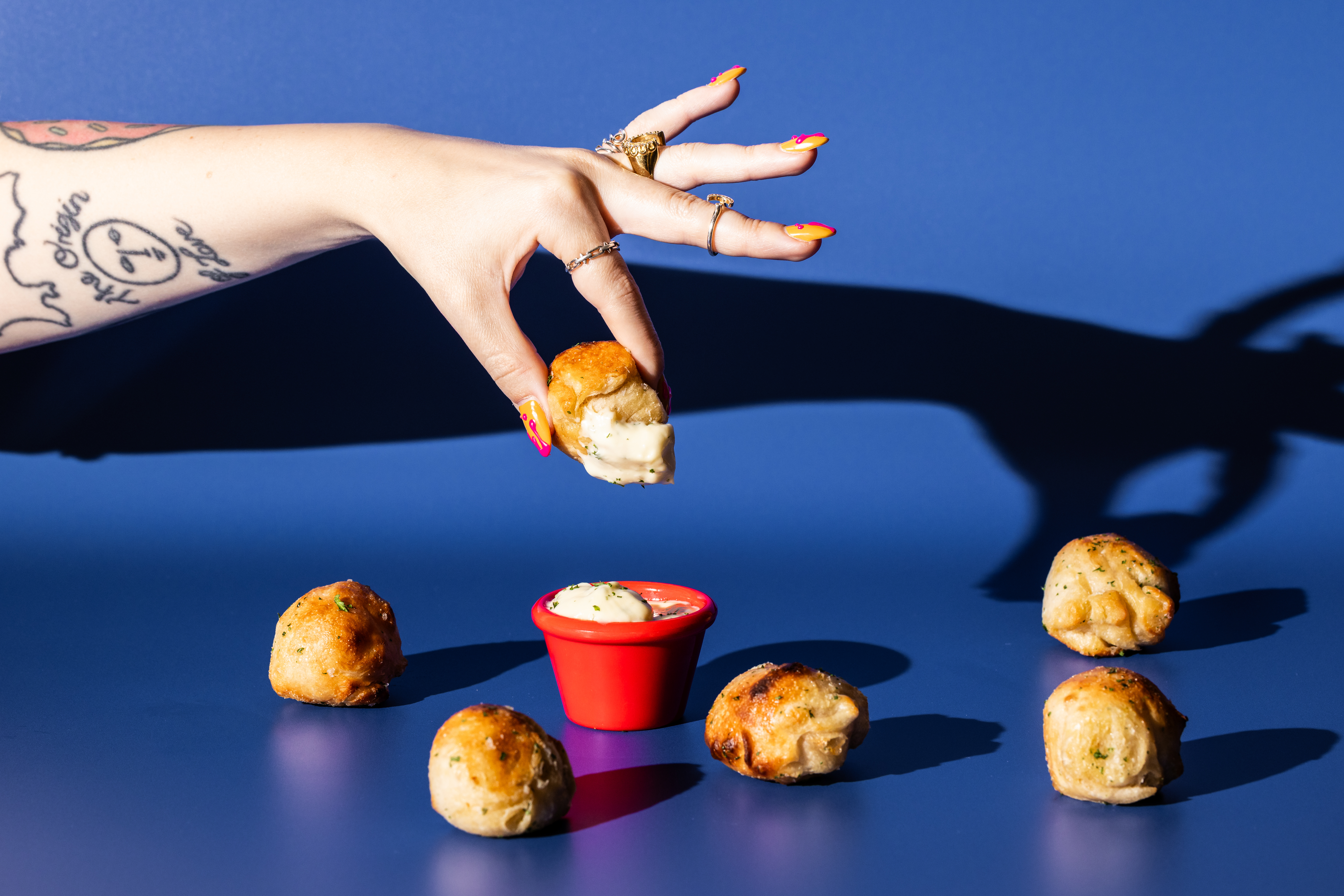 Hand dipping a garlic dough bite into creamy sauce, with more dough bites on a blue background.