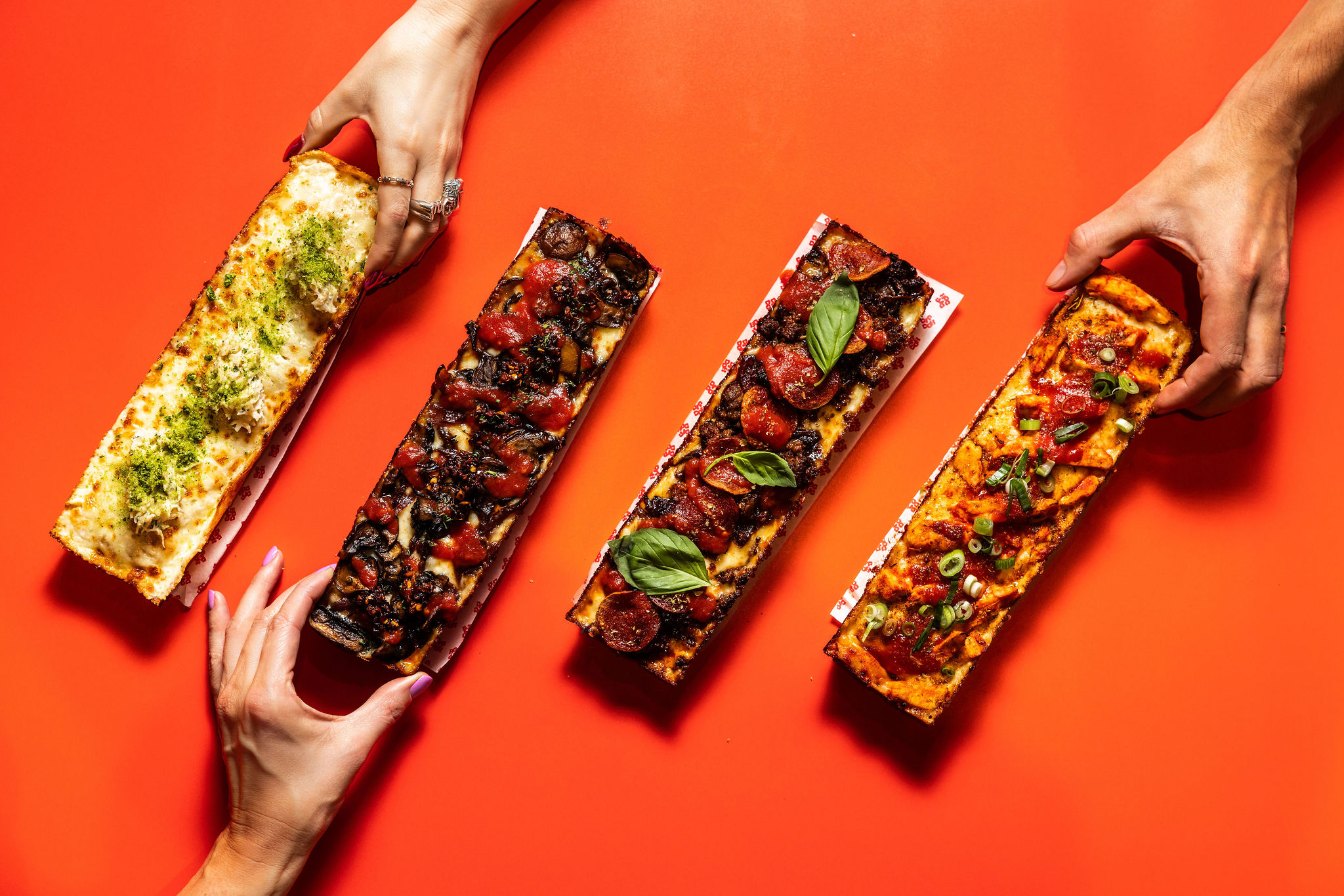 Four Detroit-style pizzas with assorted toppings on an orange background, with hands reaching in.